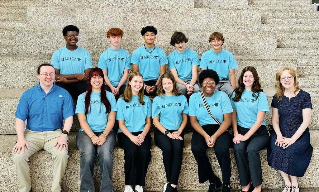 Ten students in teal T-shirts sit with two adult sponsors