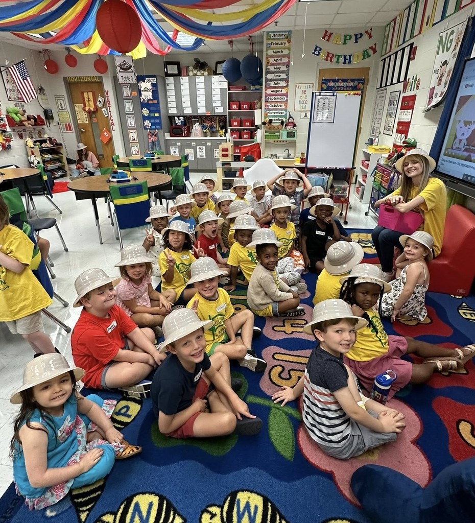 A group of students wearing plastic safari-style hats sit on a rug in front of a teacher in a classroom