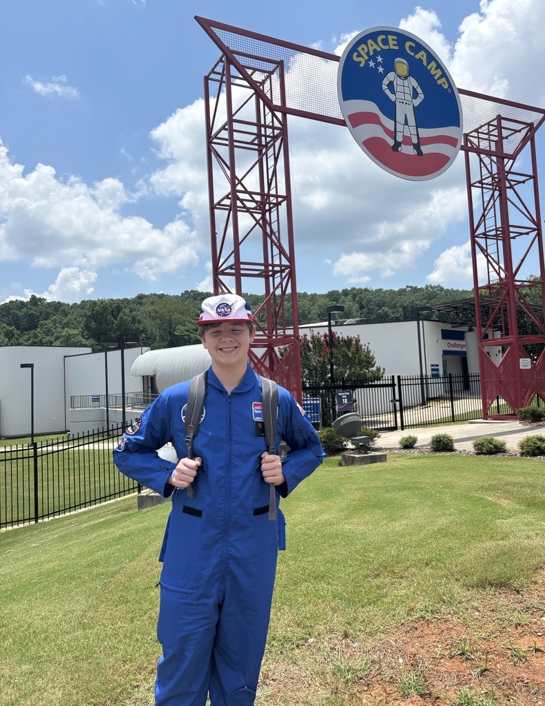 Liam Norris, dressed in a blue flight suit and white cap, stands in front of the Space Camp sign