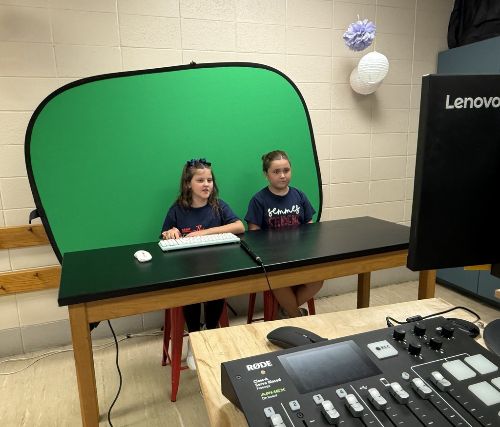 Two students sit at a table in front of a green screen, practicing morning announcements