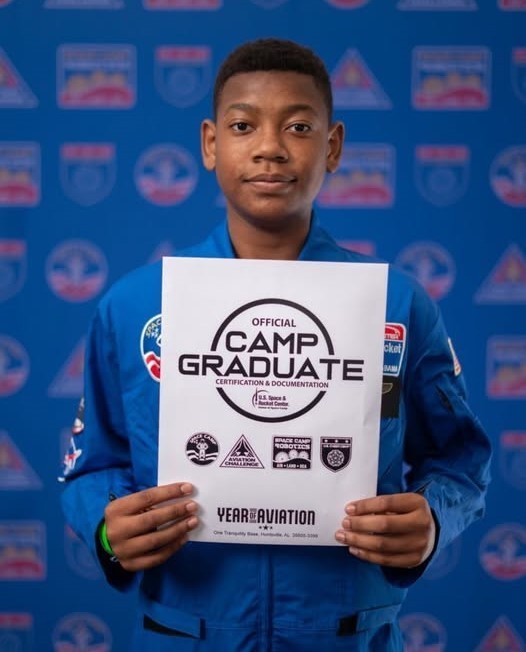A boy in a blue flight suit holding a certificate that reads "Official Camp Graduate"