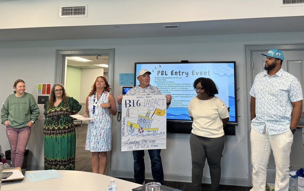 A row of teachers in a classroom, with one holding a sign that reads "Big Impact"