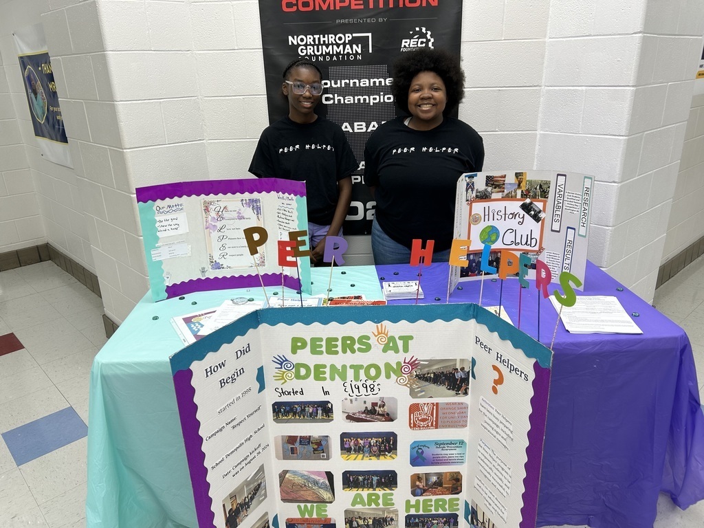 Two students stand at a table behind a poster board that reads "Peers at Denton"