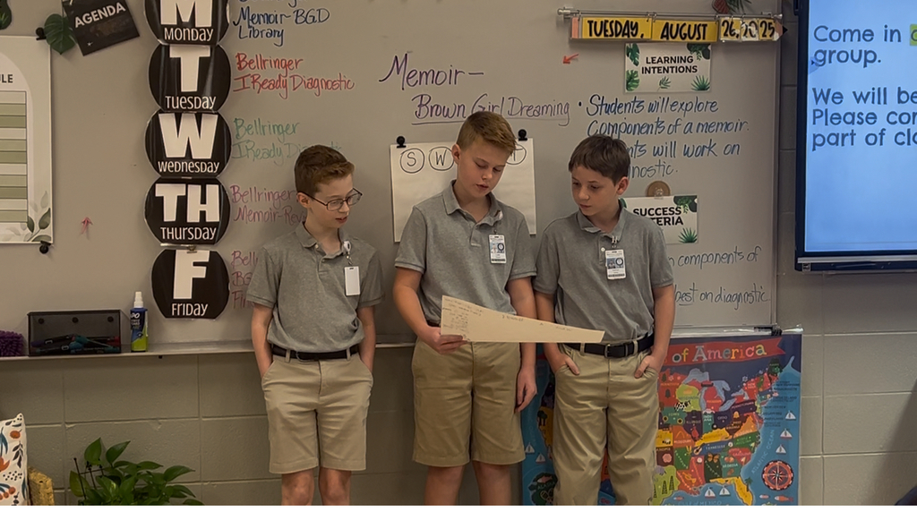 3 boys standing in front of a class, looking at a poster that 1 is holding. He is reading something from the poster.