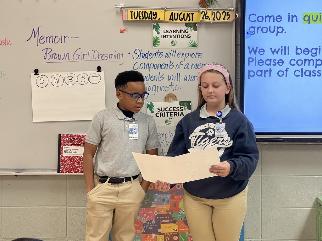 A boy and girl standing in front of a class. The girl is holding a poster. The boy is looking at it. The girl is reading something on it.