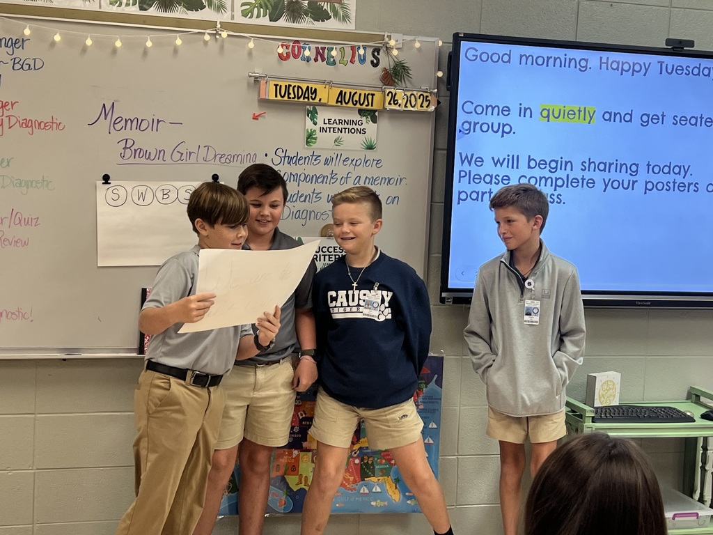 4 students standing in front of a class. 3 of them looking at a poster. One is reading from the poster.