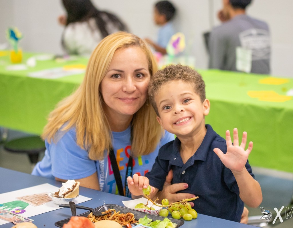 A boy smiles and waves while sitting at a cafeteria table with his grandmother