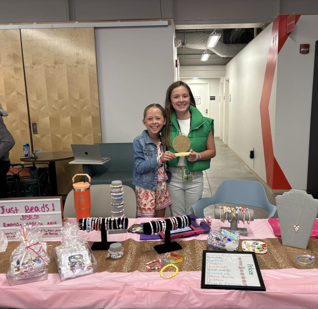 Two girls holding a trophy, standing behind a table filled with bracelets and necklaces