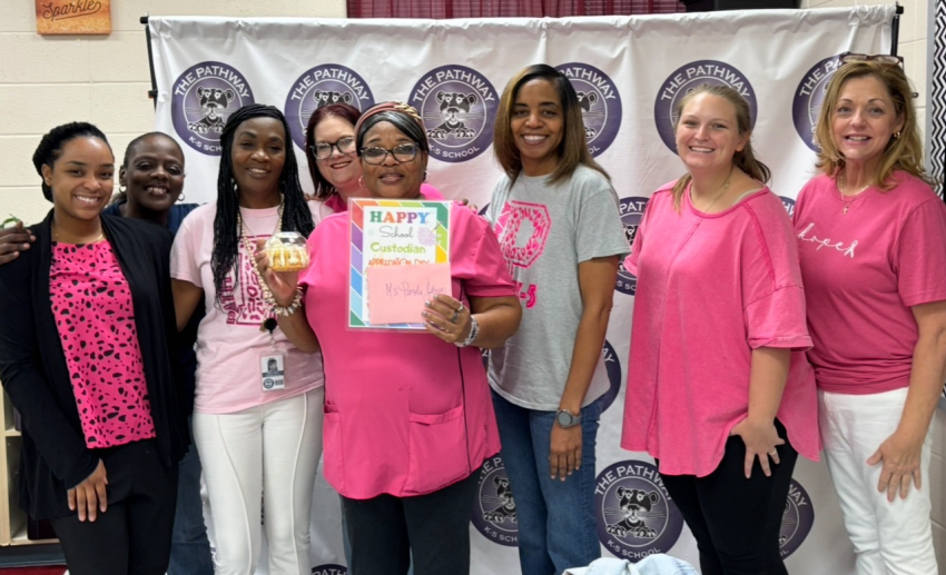 Teachers are gathered around to celebrate the custodian Ms. Johnson for national custodian day. Ms. Johnson is holding  a special card, and cake.  Everyone is dressed in pink. 