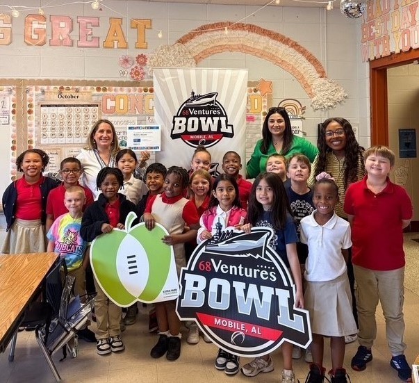A group of elementary children stand with their teacher and two other adults, with a sign from the 68 Ventures Bowl