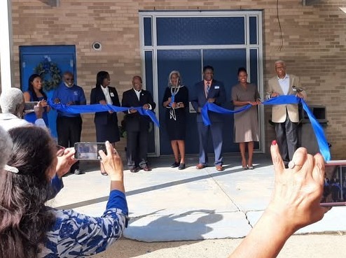 Several people cut a blue ribbon in front of a building, as people in the foreground take photos