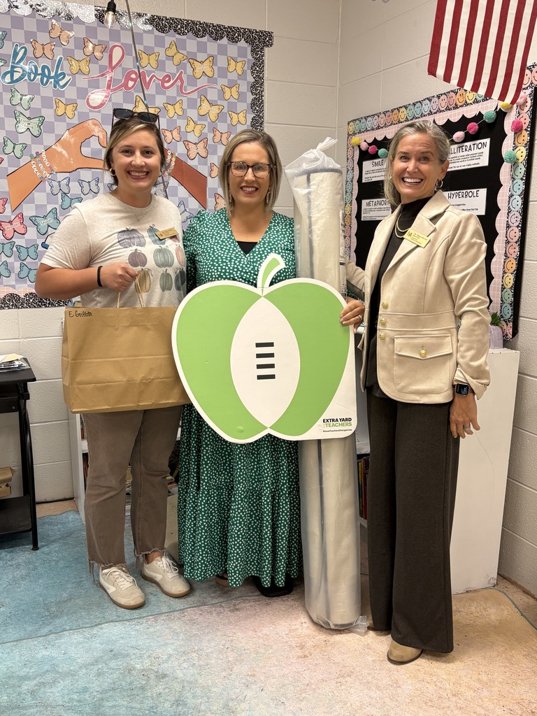 Picture of teacher and 2 other women, holding an apple sign, a bag of gifts, and a new rug