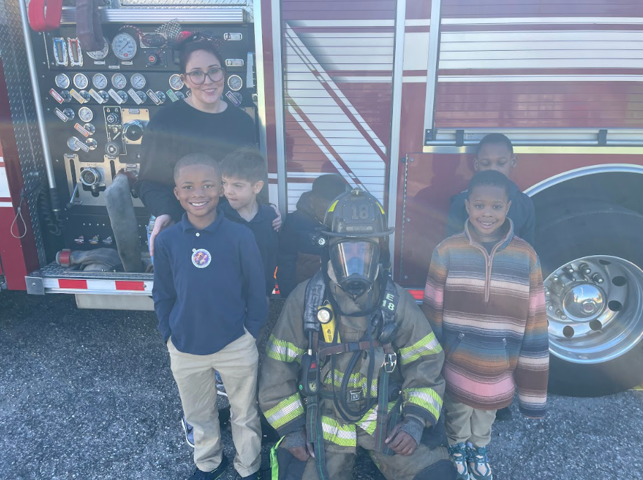 Students and their teacher standing in front of a fire truck, with a fire fighter kneeling down in front of them