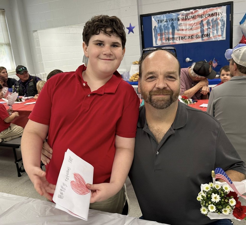 A father and son. The son is holding a hand-drawn card that reads "Happy Veterans Day"