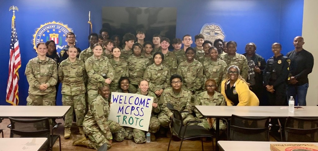 A group of JROTC cadets in fatigues, holding a sign that reads "Welcome MCPSS JROTC"