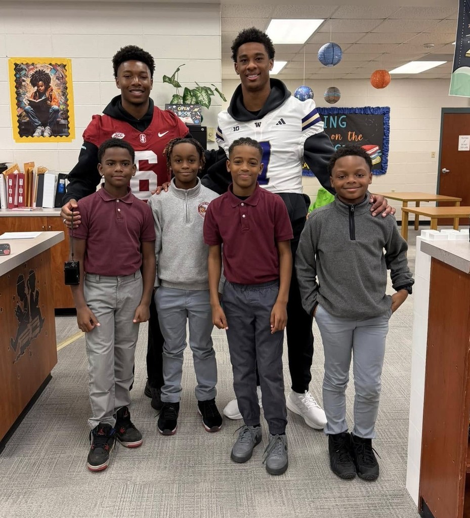 Two college football players in jerseys stand with four elementary students