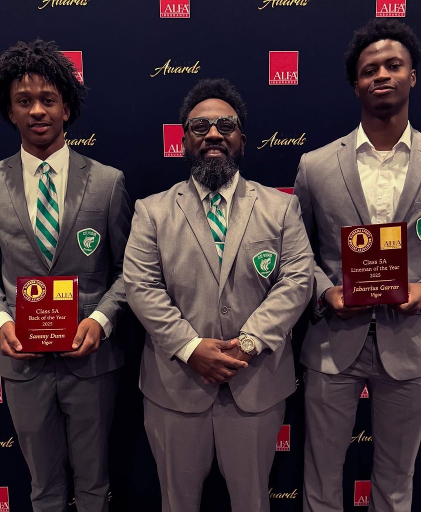 A man in a gray suit stands with two high school boys in gray suits who are holding plaques