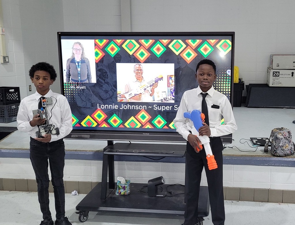 One student holds a robot and another holds a Super Soaker in front of a screen that features Lonnie Johnson