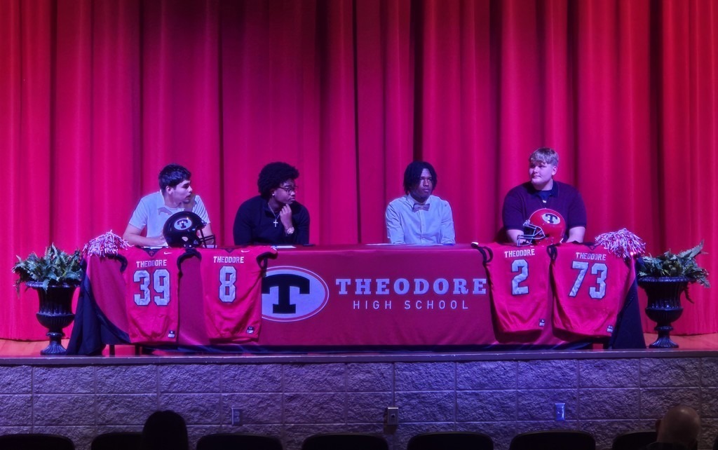 Four high school boys sit on a stage behind a table that reads "Theodore High School"