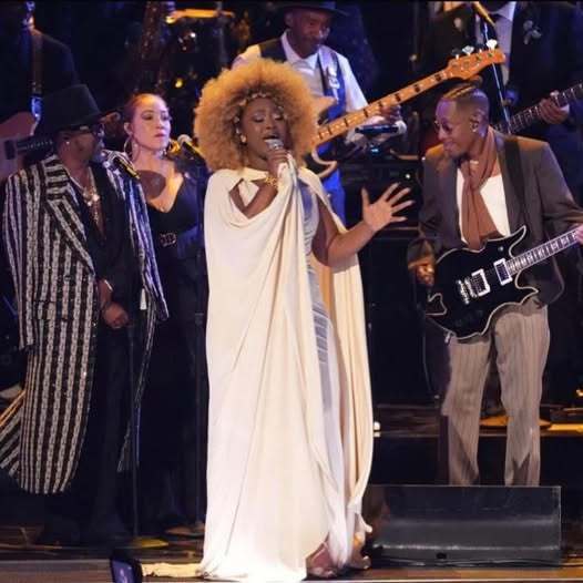 A woman in a white gown signing on a stage, surrounded by other musicians