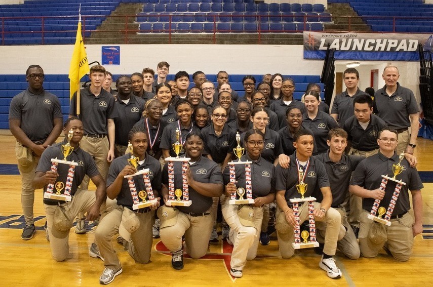 A large group of students in gray shirts and khaki pants, with several in the front holding trophies