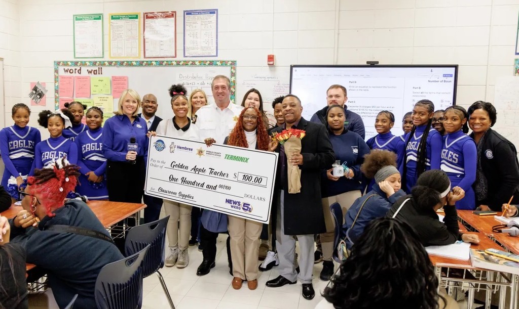 A teacher holding a large check in a classroom, surrounded by cheerleaders and a few adults