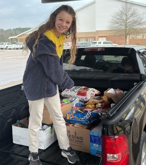 A girl stands in the bed of a pickup truck, pointing at bags of dog food in it