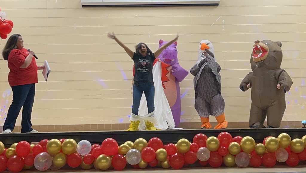 A teacher stands on a stage with her arms raised,  in front of others in inflatable costumes