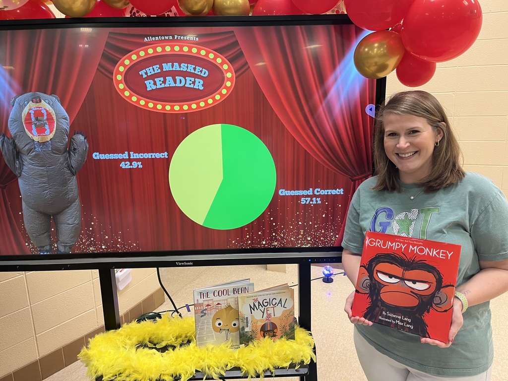 A teacher holding a book in front of a screen that shows her dressed in an inflatable costume