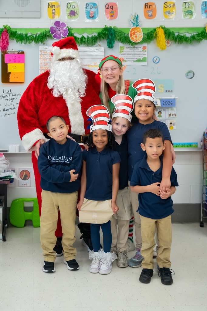 A man in a Santa suit stands with a teacher in an elf costume and several children