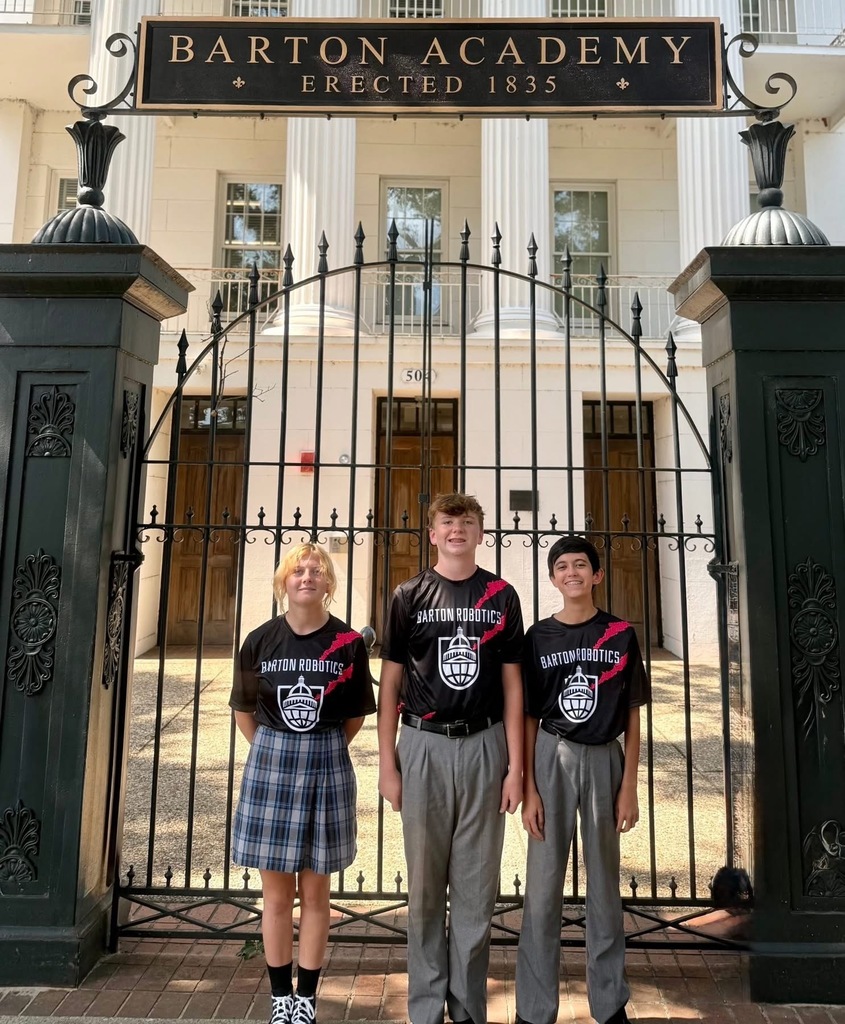 Three students wearing "Barton Robotics" T-shirts, standing outside the front gate of the school