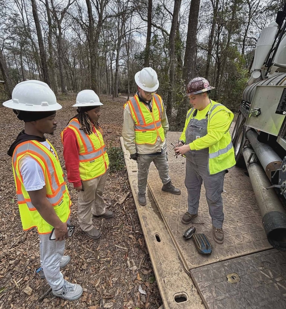 A man in a hard hat talking with three Theodore High students in hard hats in the woods.