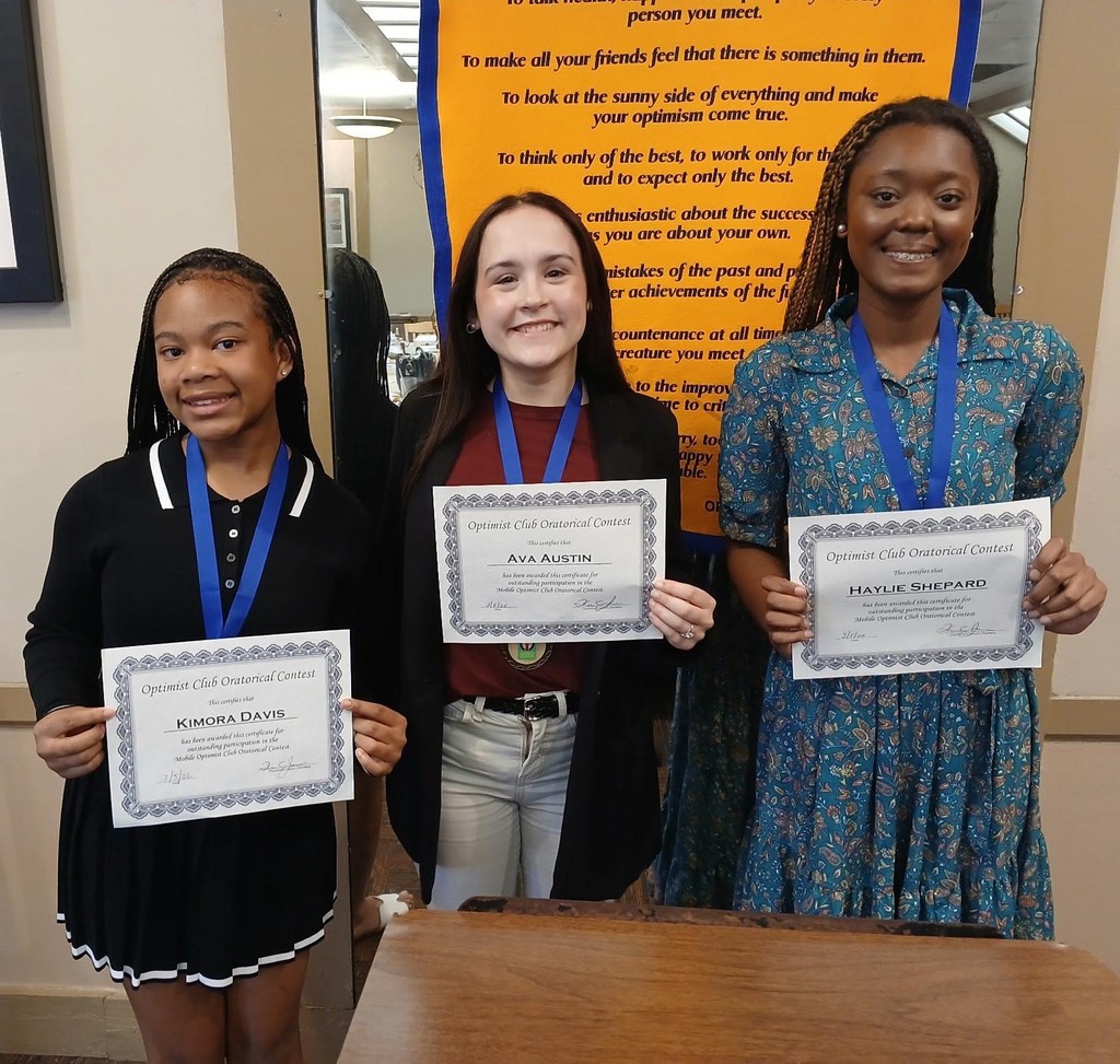 Three girls holding certificates in front of a banner with the Optimists Creed