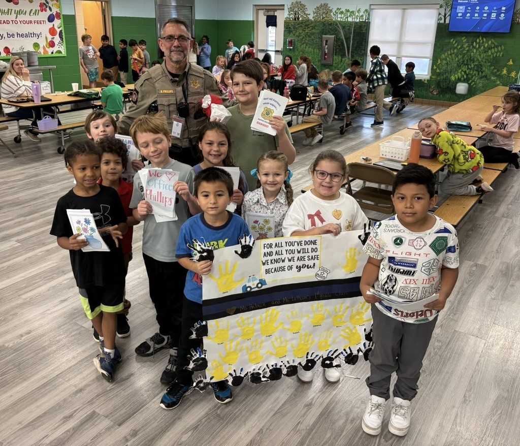 A group of Wilmer Elementary students stand in a cafeteria with a sheriff's deputy and a sign thanking him