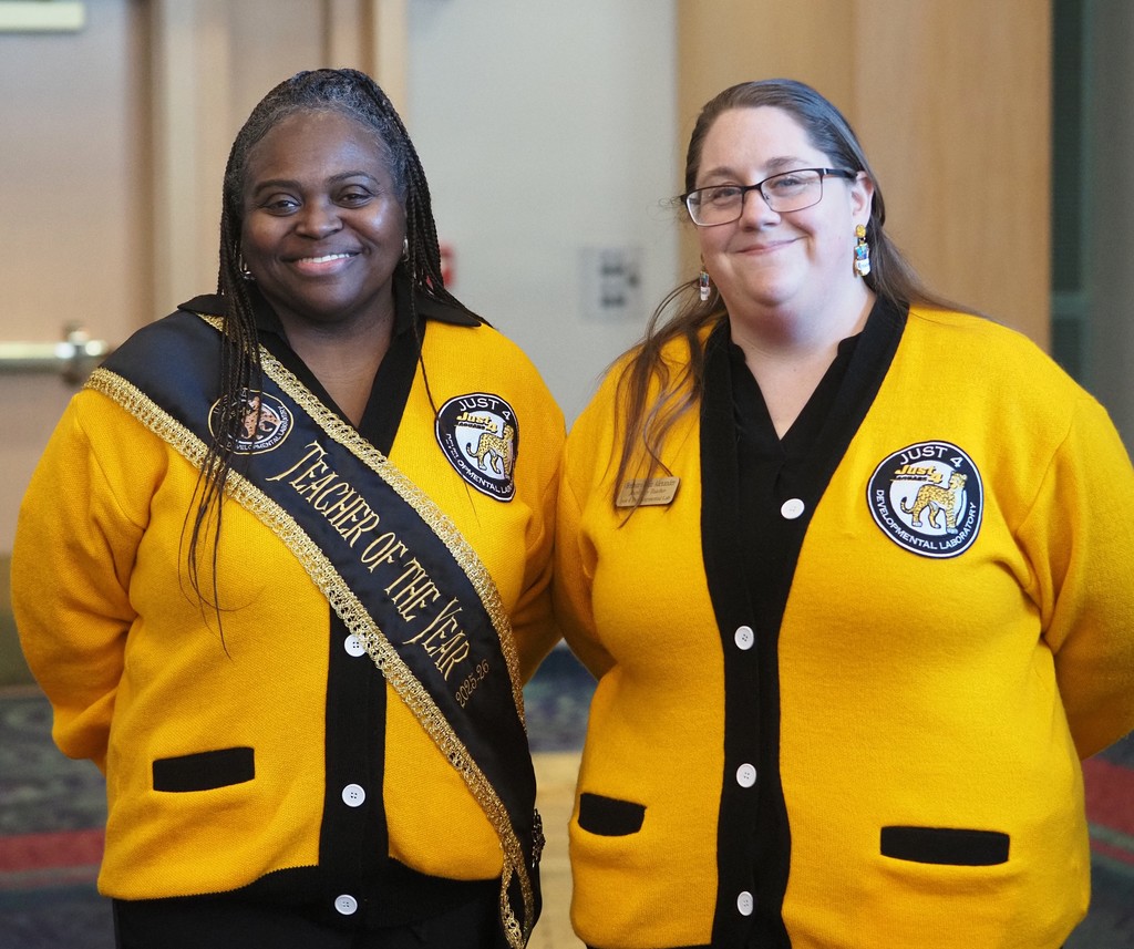 Two representatives of Just 4 in yellow sweaters, with one wearing a Teacher of the Year sash
