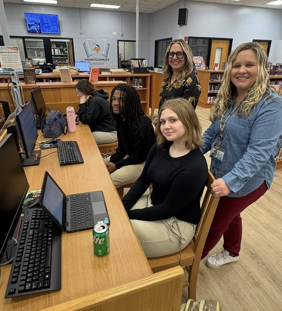 Two high school students sit at computers with two teachers standing behind them