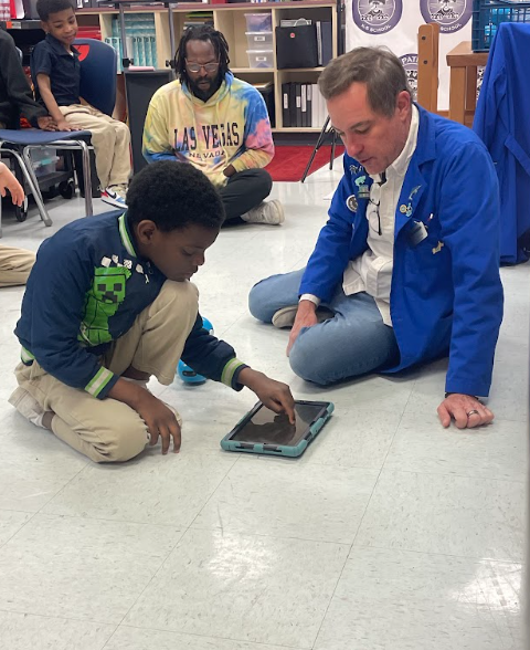 A student sitting on the floor next to an adult. The adult is teaching the student how to program a tablet to make a robot move 