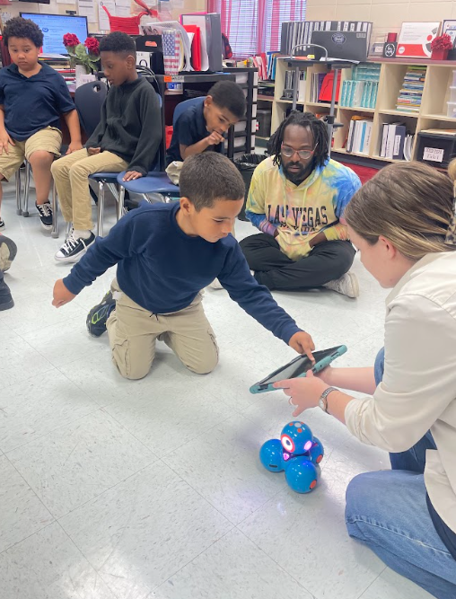 A student sitting on the floor next to an adult. The adult is teaching the student how to program a tablet to make a robot move 