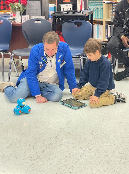 A student sitting on the floor next to an adult. The adult is teaching the student how to program a tablet to make a robot move 