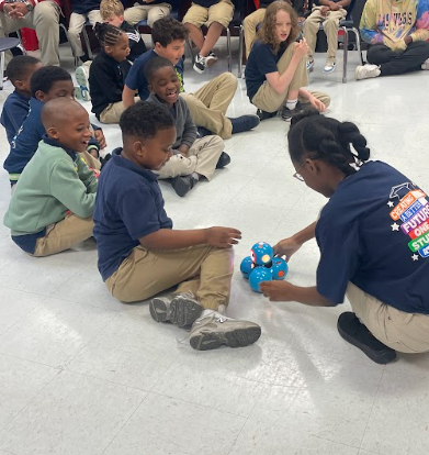 Students seating on the floor gather around as they watch a robot maneuver across the room 
