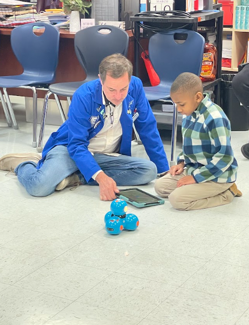 A student sititng on the floor next to an adult. The adult is teaching the student how to program a tablet to make a robot move 