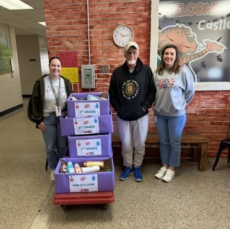 Three adults standing with boxes of toiletries, separated by grade level
