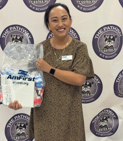 **ALT Text:** A smiling woman stands in front of a step-and-repeat banner with “The Pathway K-5 School” logos. She is holding a clear gift bag filled with school or hygiene supplies, including bottles and packaged items labeled “AmFirst.” She wears a brown patterned dress, a name badge, and a smartwatch, and appears to be posing at a school-related event.