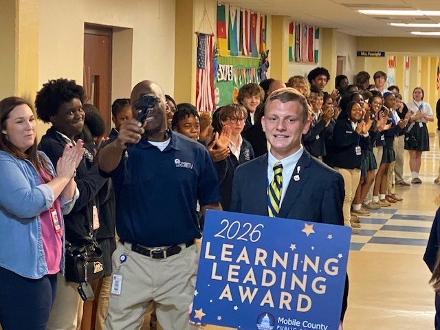 Students in a hallway clap for Ben Stringfellow as he carries a Learning Leading Award sign