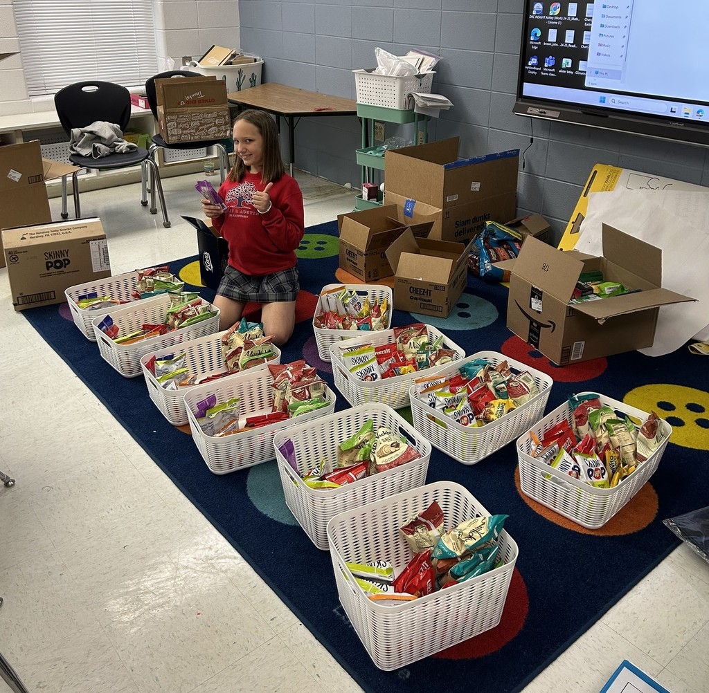 A girl giving a thumbs up, surrounded by baskets of snacks on a classroom floor