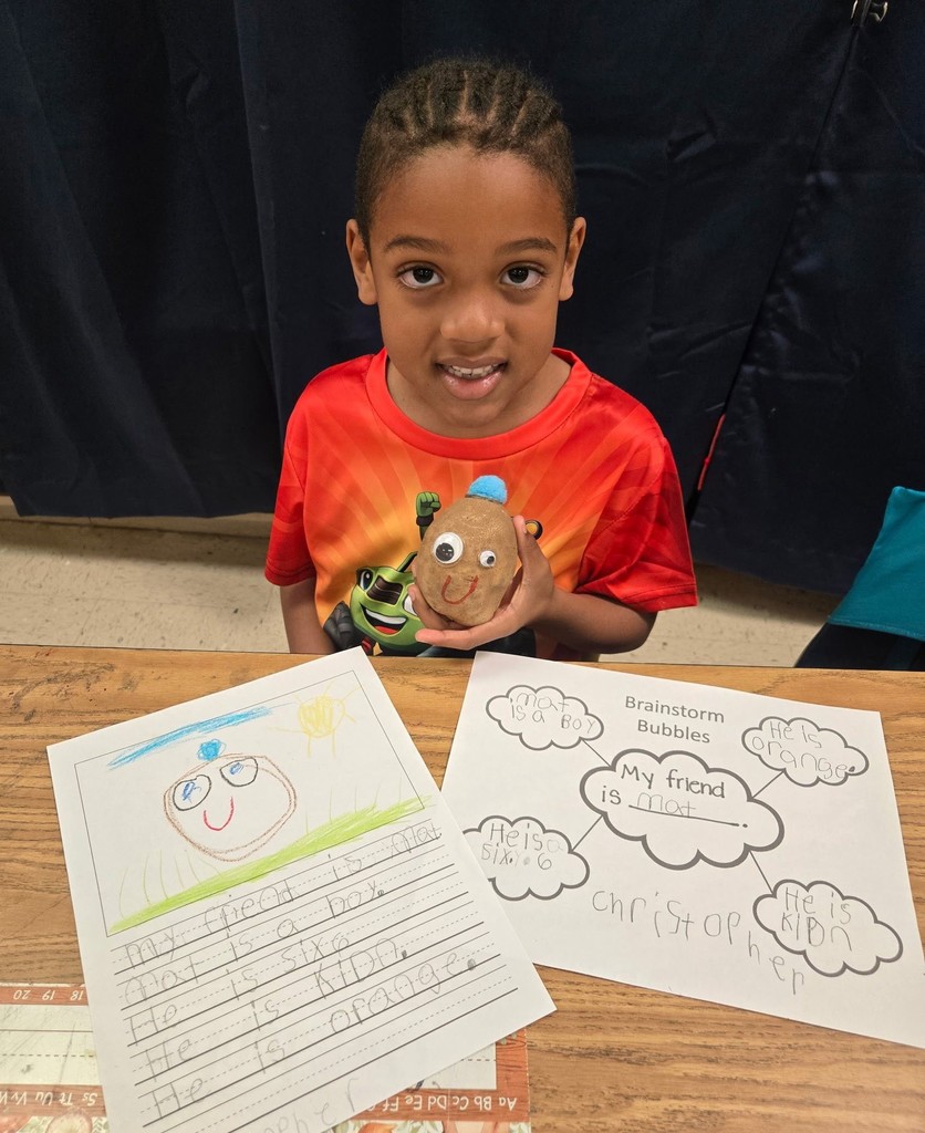 A boy holds a potato with a smiley face on it, with worksheets on the table in front of him