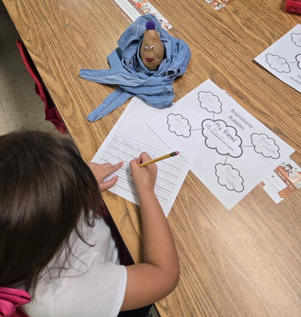 A girl writes on a worksheet, with a potato swaddled in a blanket on the table near her