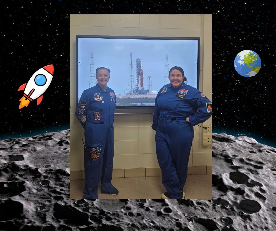 Two Davidson High teachers in astronaut suits stand in front of a screen showing a rocket launch