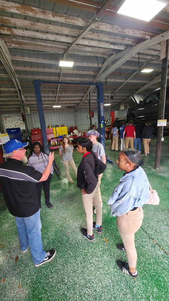 A group of students touring a mechanics garage with a car on a lift.