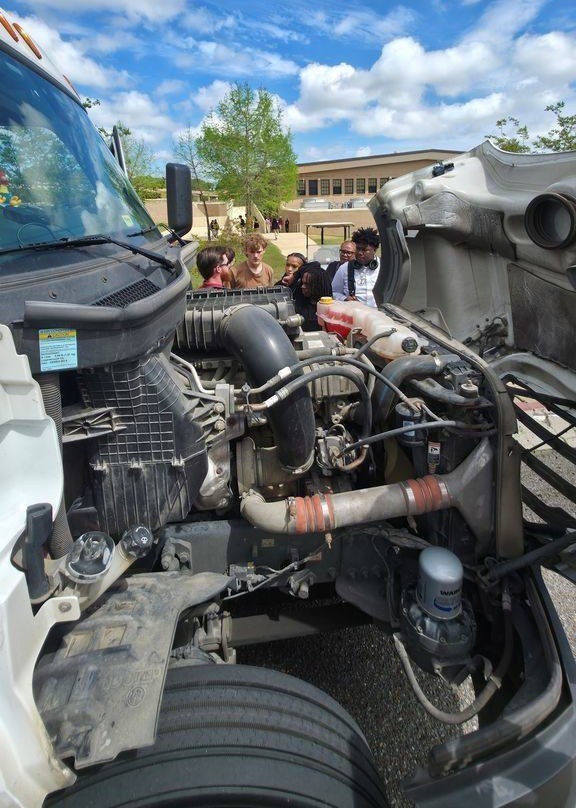 A man talks to Theodore High students with the exposed engine of a semi truck in the foreground